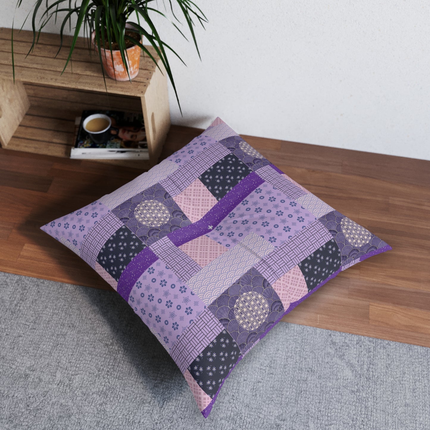 A square patchwork floor pillow in shades of purple, lavender, and dark gray with floral and geometric patterns, styled on a wooden bench in a room setting.