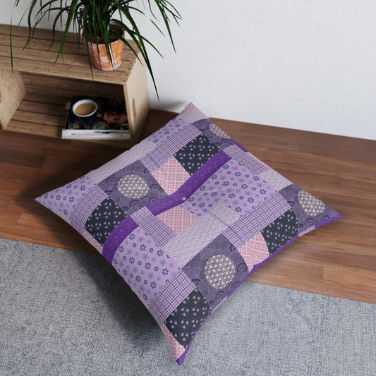 A square patchwork floor pillow in shades of purple, lavender, and dark gray with floral and geometric patterns, styled on a wooden bench in a room setting.