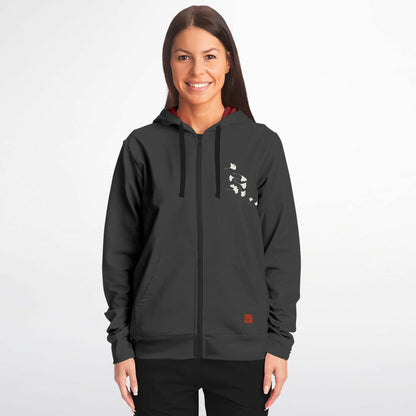 A woman modeling a dark gray zip-up hoodie with a small white graphic on the left chest and a small brown square patch near the lower right hem, set against a plain light gray background.