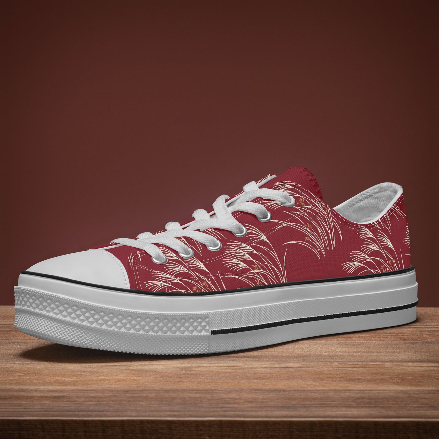 A low-top canvas sneaker with a red upper featuring a white palm leaf pattern, white laces, a white rubber toe cap, and a thick white platform sole with a black stripe, resting on a wooden surface against a solid dark brown background.