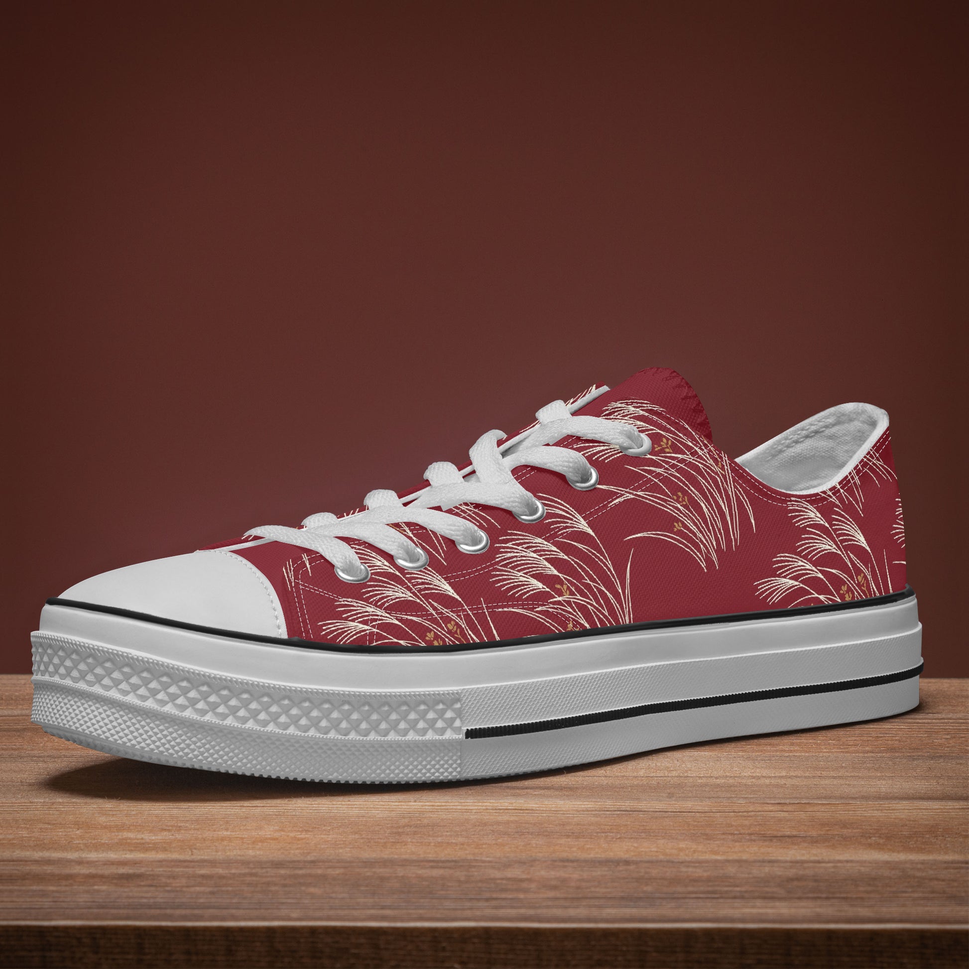 A low-top canvas sneaker with a red upper featuring a white palm leaf pattern, white laces, a white rubber toe cap, and a thick white platform sole with a black stripe, resting on a wooden surface against a solid dark brown background.