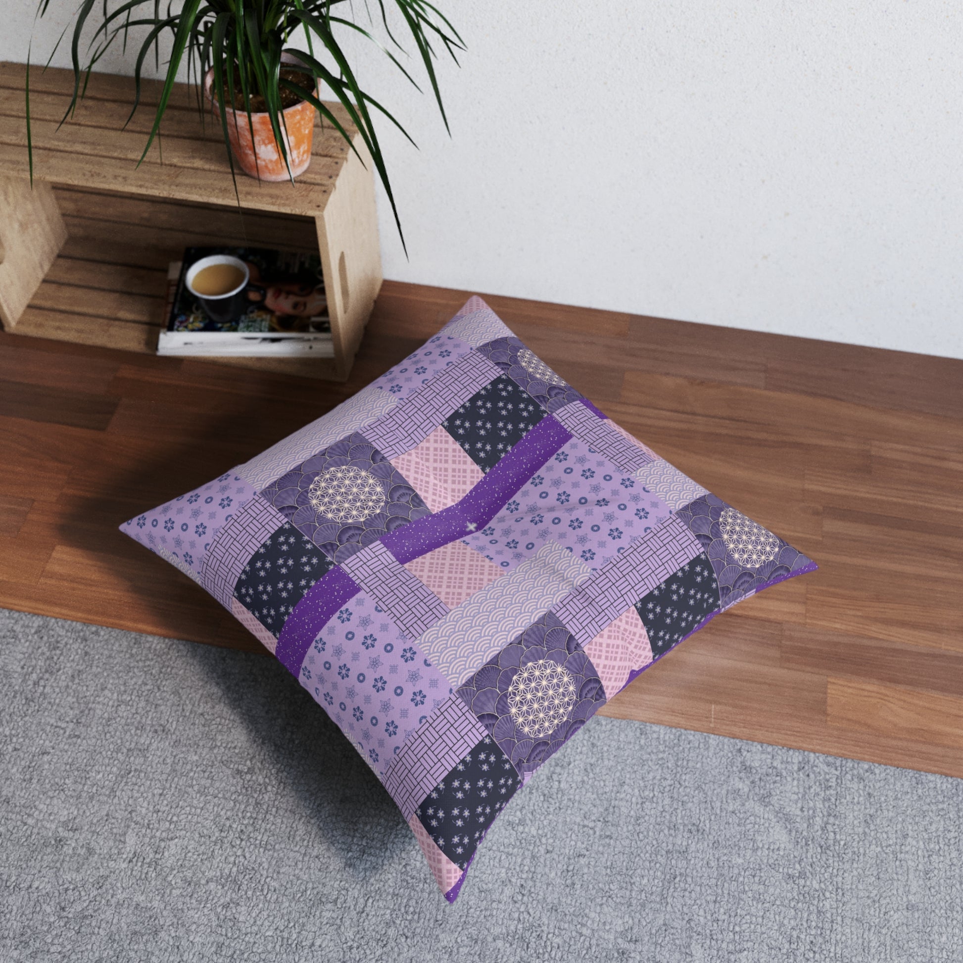A square floor cushion with a purple and lavender patchwork quilted design, styled on a gray rug next to a wooden shelf with a potted plant.