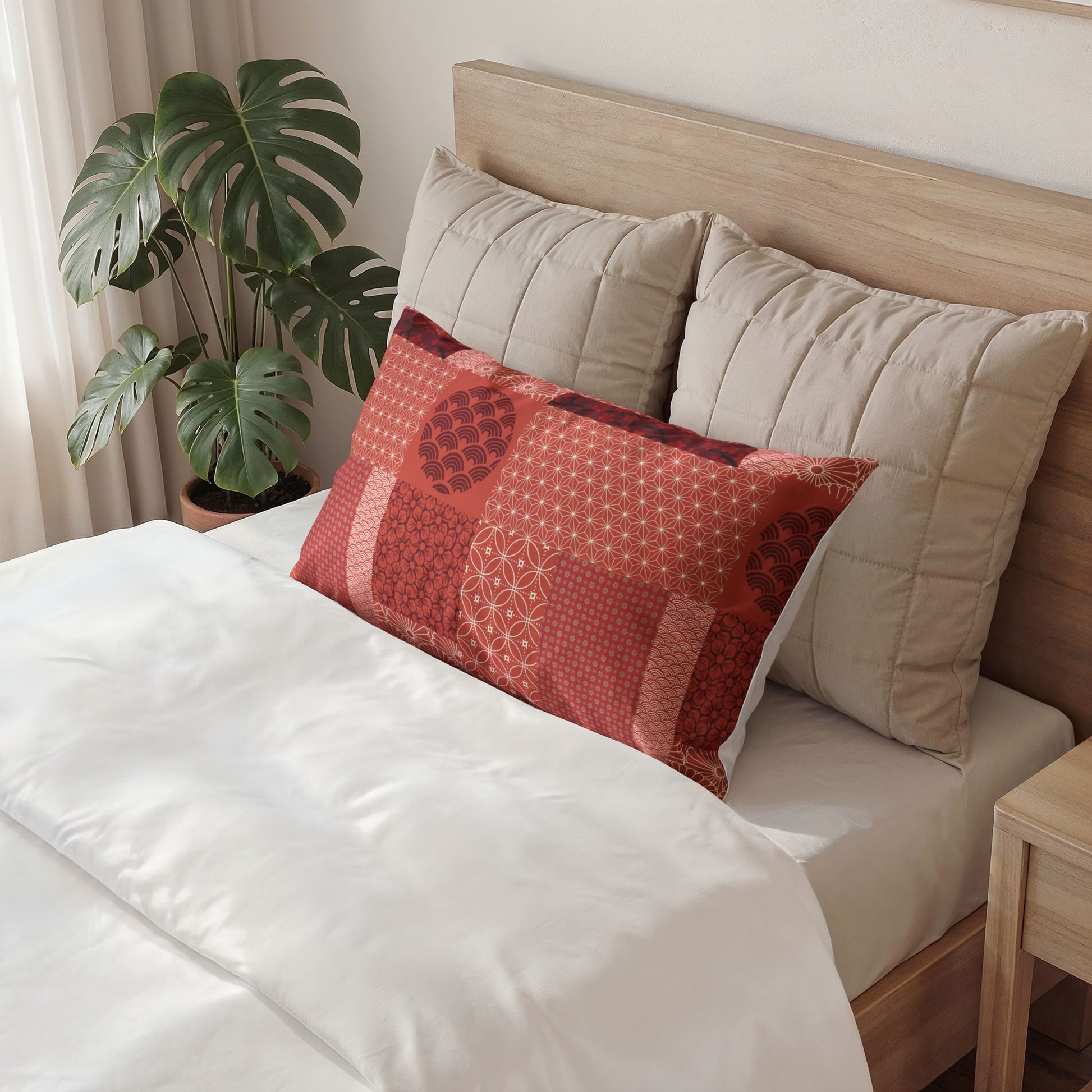 A red and orange patchwork-patterned decorative pillow resting on a bed with white bedding and two beige pillows, set against a light wood headboard with a potted green plant in the background.