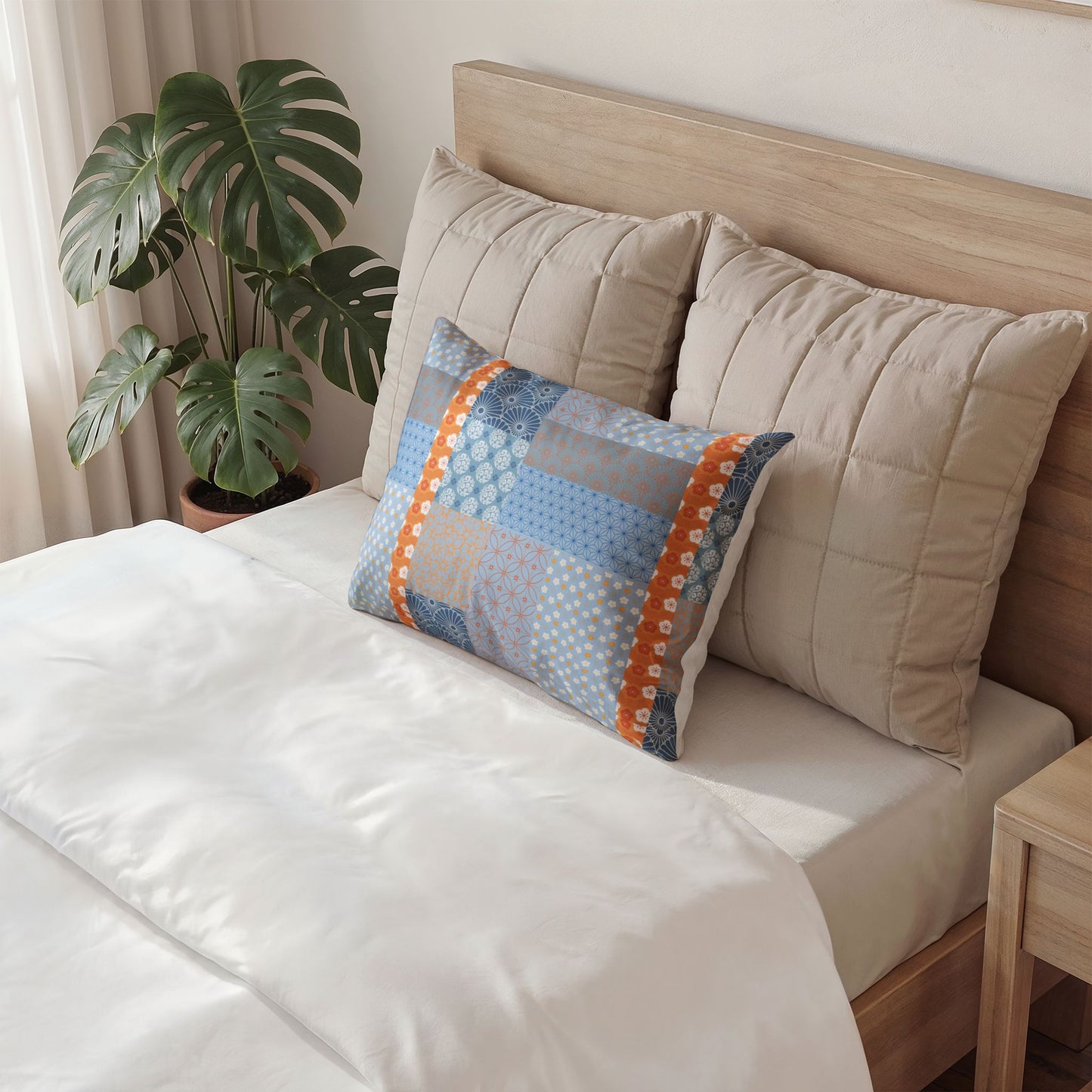 A styled bedroom scene showing a bed with white bedding, two beige quilted pillows, and a smaller rectangular patchwork accent pillow in shades of blue, gray, and orange. A wooden headboard, plant, and bedside table are visible.