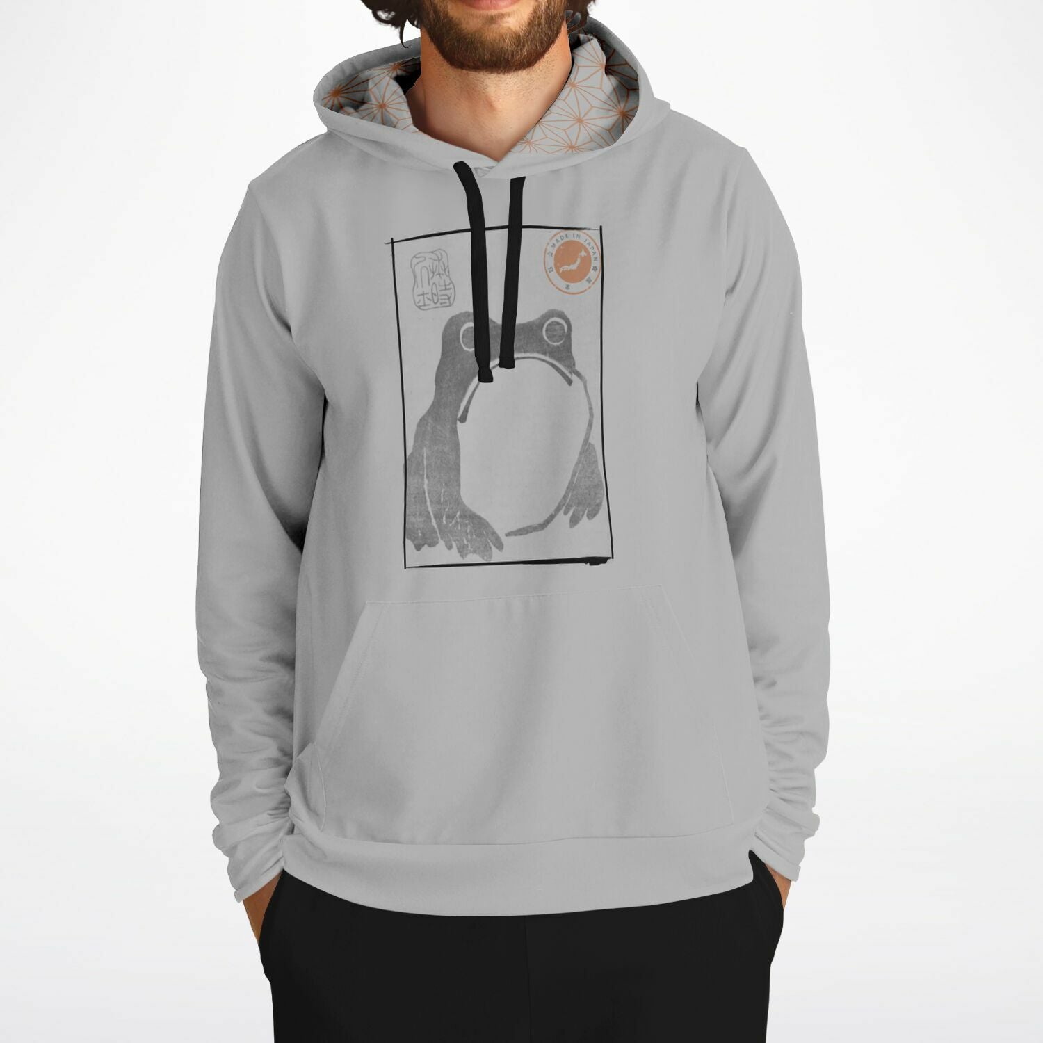 A man wearing a light gray pullover hoodie with a black and white graphic print of a frog inside a square frame on the chest, paired with black pants, against a plain white background.