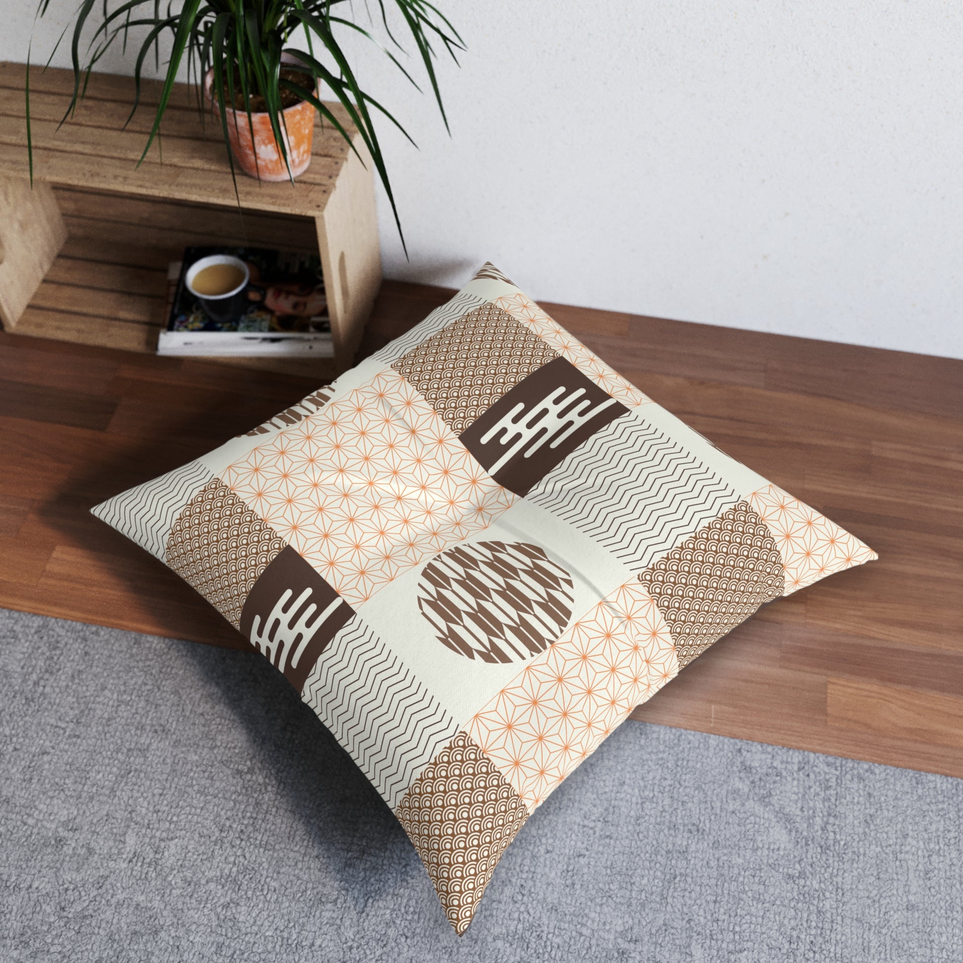A square decorative throw pillow with a patchwork of beige, brown, and orange geometric patterns, resting on a gray rug next to a wooden shelf with a potted plant.