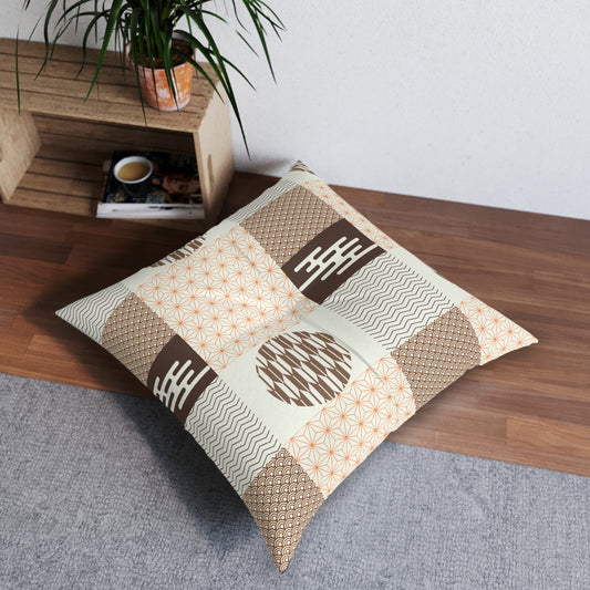 A square decorative throw pillow with a patchwork of beige, brown, and orange geometric patterns, resting on a gray rug next to a wooden shelf with a potted plant.