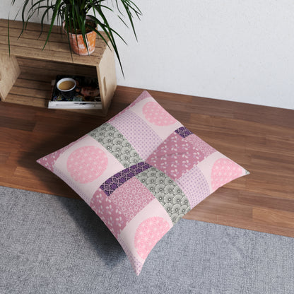 A square decorative throw pillow with a patchwork design of pink, purple, and green patterns, resting on a gray rug next to a wooden crate in a room setting.