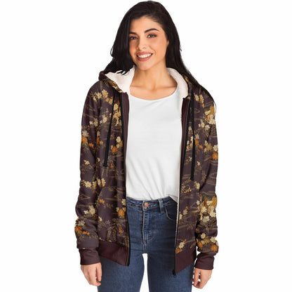 A woman modeling a brown hooded zip-up jacket with an all-over floral pattern in muted gold, beige, and olive tones, worn over a white t-shirt and blue jeans against a plain white background.