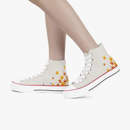 A pair of high-top canvas sneakers in off-white, decorated with a yellow and orange autumn leaf pattern on the sides, shown on a model's feet against a light gray background.