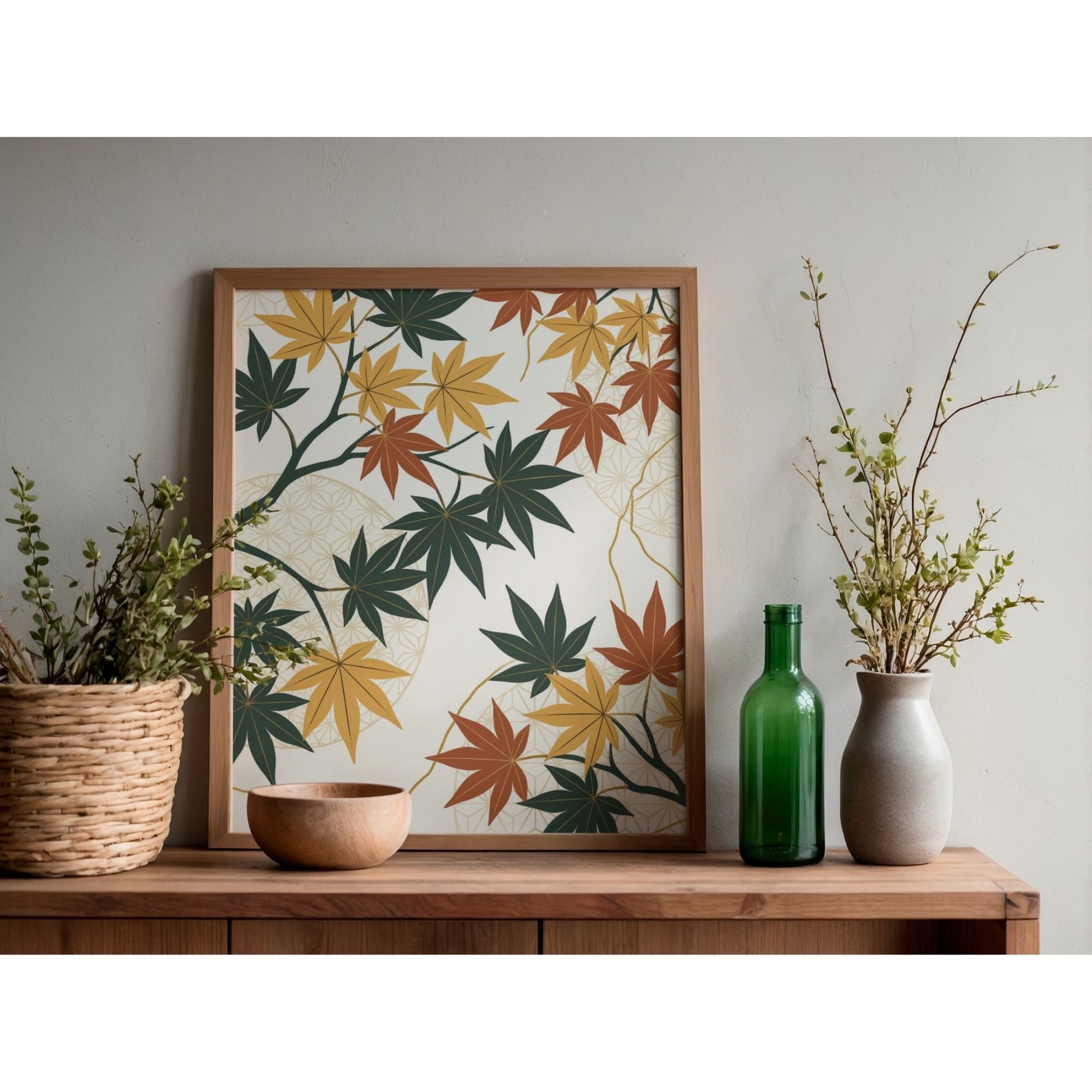 A framed art print featuring a pattern of maple leaves in green, yellow, and orange on a cream background, styled on a wooden console table with decorative plants and vases against a neutral gray wall.