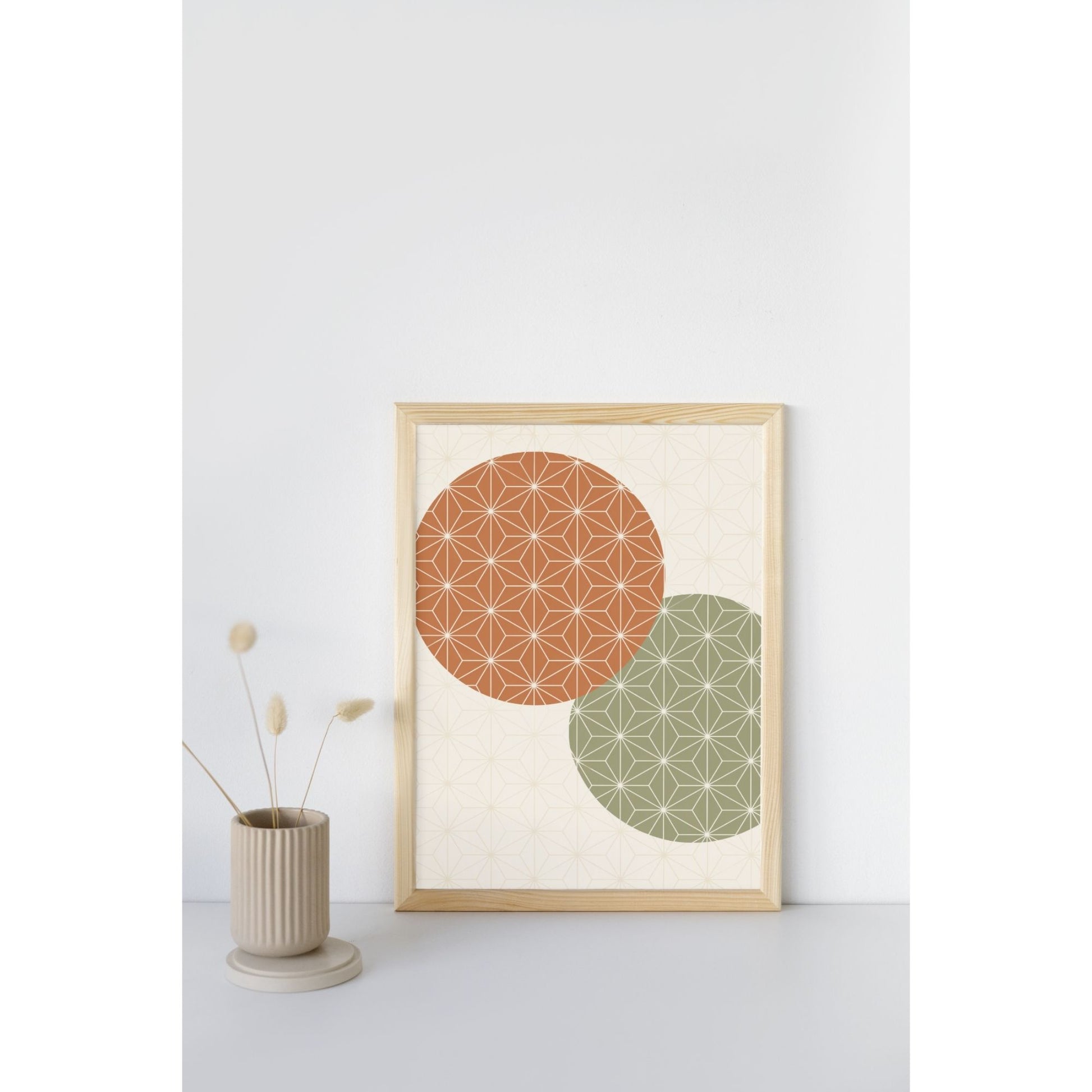 A framed art print featuring two overlapping circles with geometric star patterns, one terracotta orange and one sage green, displayed on a light gray surface next to a small beige vase with dried grass.
