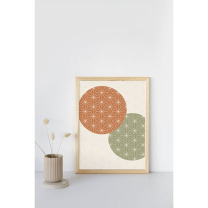 A framed art print featuring two overlapping circles with geometric star patterns, one terracotta orange and one sage green, displayed on a light gray surface next to a small beige vase with dried grass.