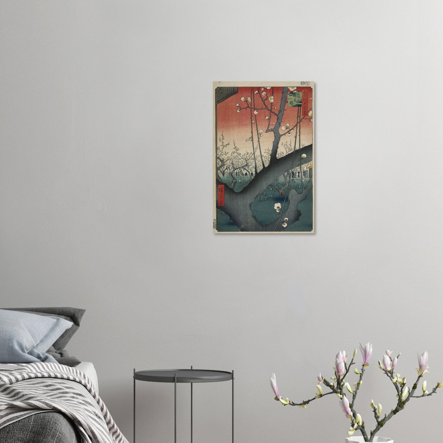 Framed artwork on a wall above a bed with a side table and plant in the foreground.