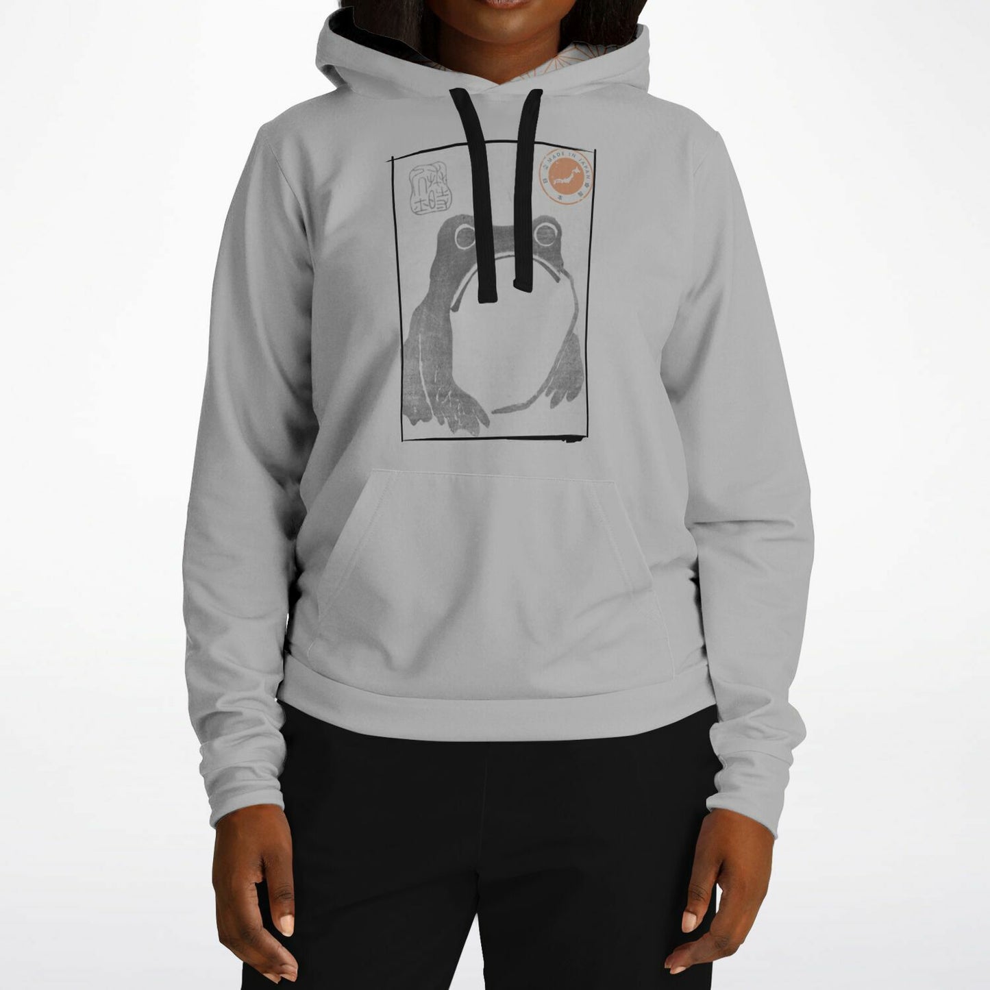 A person wearing a light gray pullover hoodie with a black drawstring and a large graphic print of a frog on the chest, paired with black pants, against a plain white studio background.
