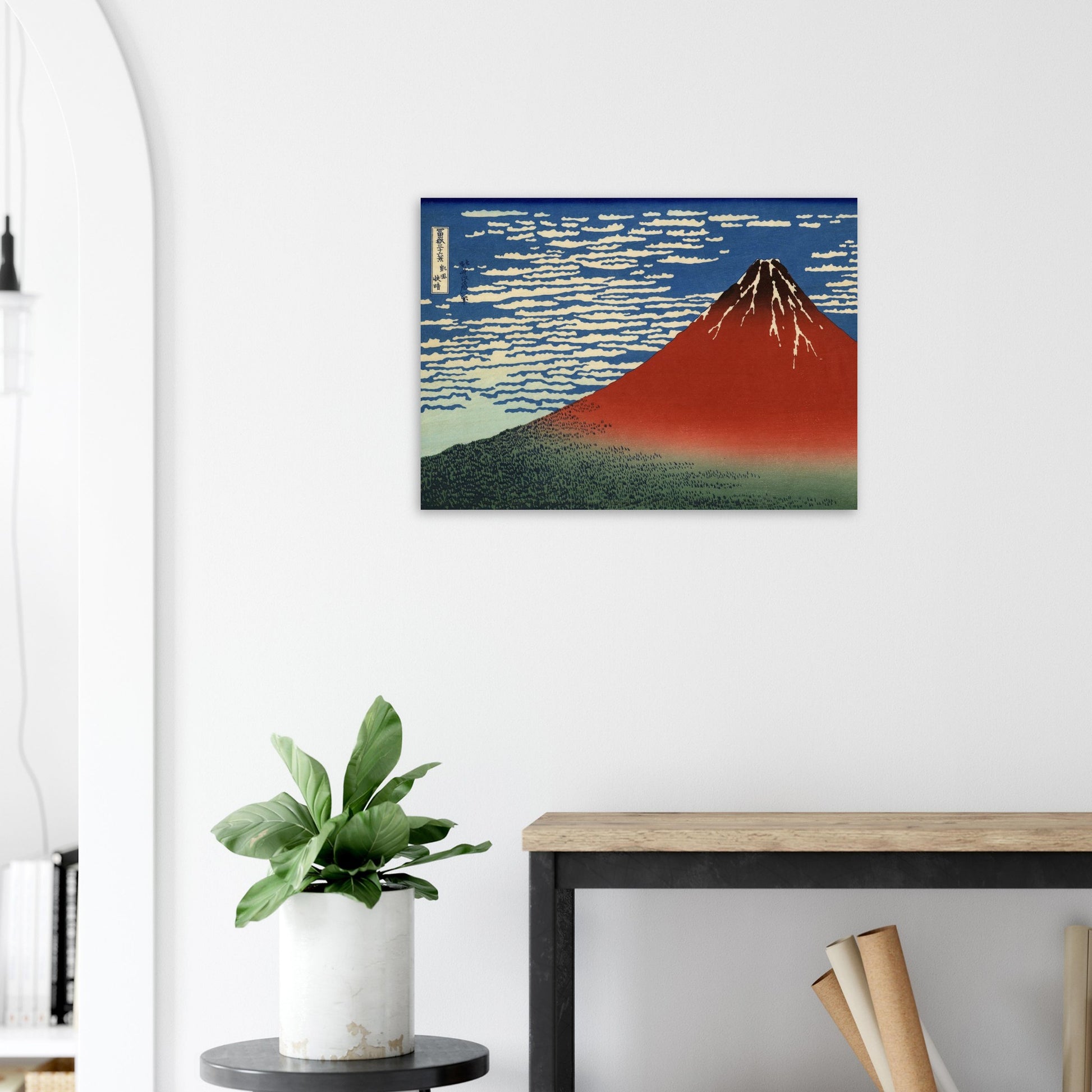 Framed artwork of a mountain on a white wall above a desk with a plant.