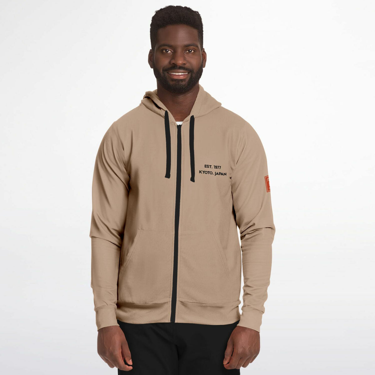 A man wearing a beige zip-up hoodie with black drawstrings, black text on the left chest reading 'EST. 2017 KYOYO JAPAN', and a small orange patch on the sleeve, photographed against a light gray studio background.