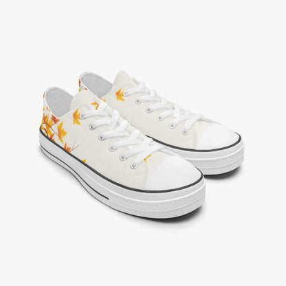 A pair of low-top canvas sneakers in off-white, decorated with a yellow-orange maple leaf pattern on the side and a black rubber trim around the toe and sole.