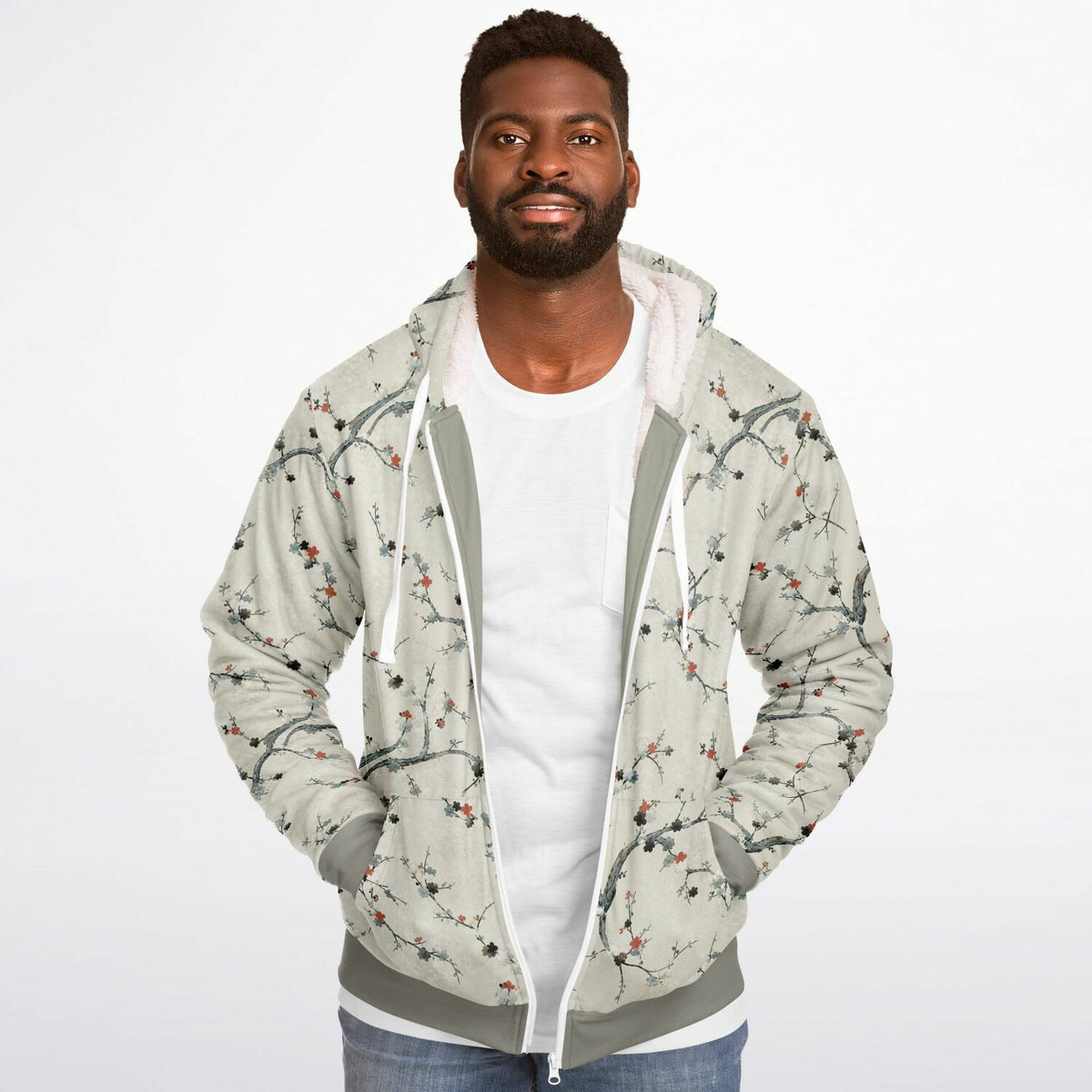 A man wearing a light beige zip-up hoodie with a delicate branch and berry print, layered over a white t-shirt and blue jeans, photographed against a seamless white studio background.