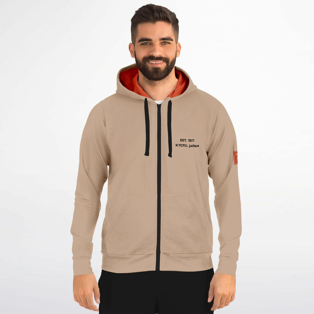 A man wearing a beige zip-up hoodie with a black front zipper, black drawstrings, a black printed logo on the left chest, and a bright orange inner hood lining, set against a plain light gray background.