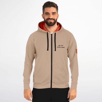 A man wearing a beige zip-up hoodie with a black front zipper, black drawstrings, a black printed logo on the left chest, and a bright orange inner hood lining, set against a plain light gray background.