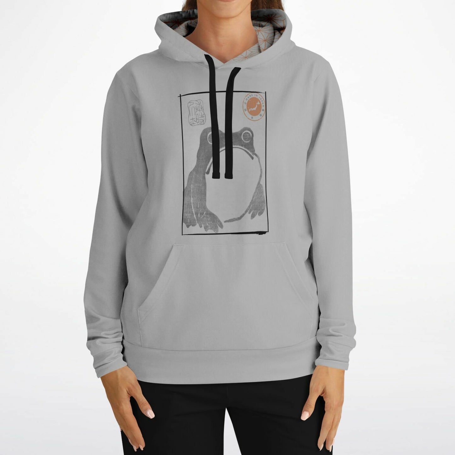 A woman wearing a light gray pullover hoodie with a black and white graphic print of a frog on the chest, paired with black pants, against a plain white background.