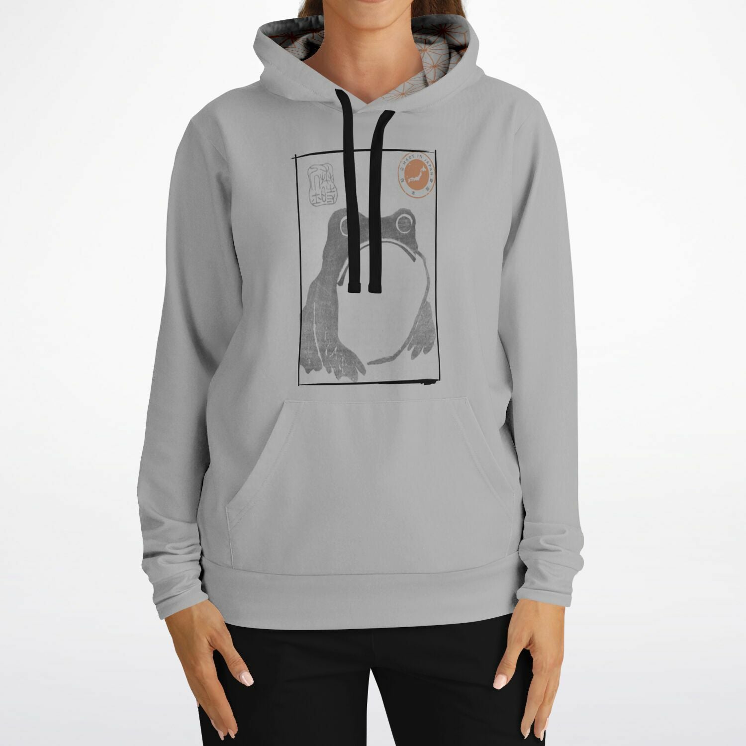 A woman wearing a light gray pullover hoodie with a black and white graphic print of a frog on the chest, paired with black pants, against a plain white background.