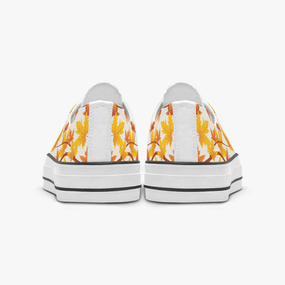 A pair of low-top canvas sneakers shown from the back, featuring a white background with a yellow and orange autumn leaf pattern on the sides and thick white platform soles with black stripes.