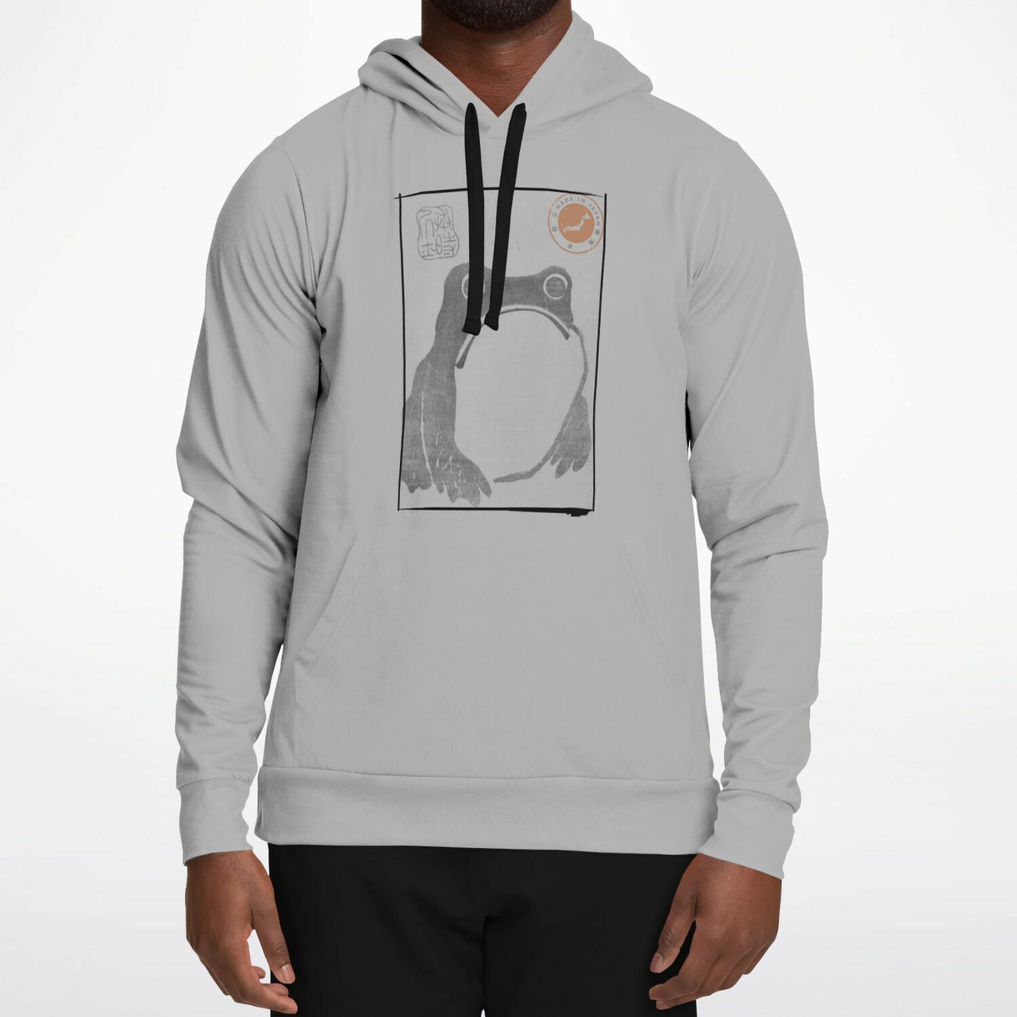A man wearing a light gray pullover hoodie with a black and white graphic print of a frog on the chest, paired with black pants, against a plain white background.