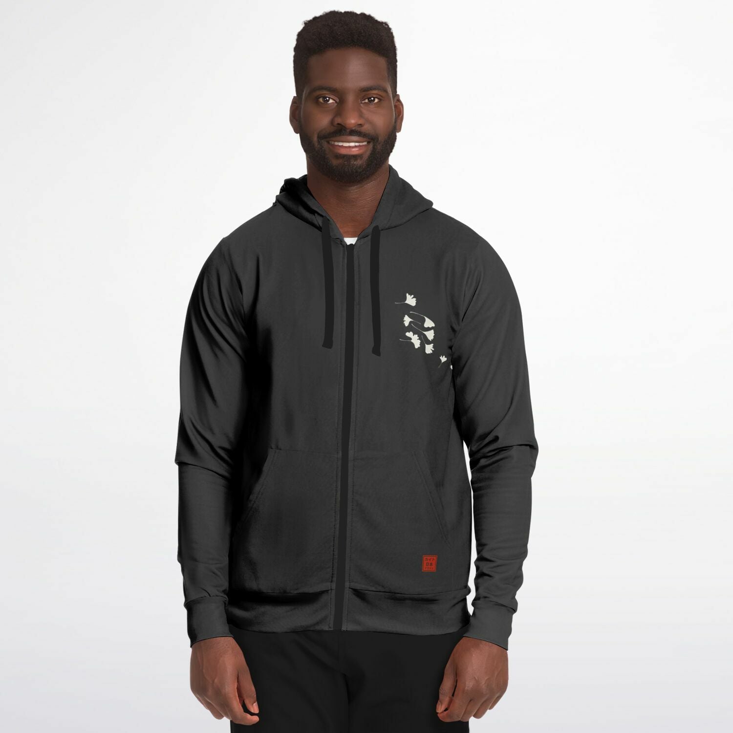 A man wearing a black zip-up hoodie with a small white graphic on the left chest and a small red square logo near the lower hem, photographed against a light gray studio background.